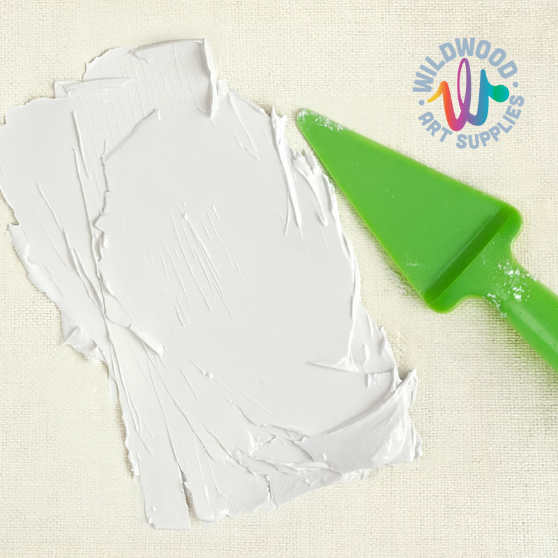 A thick, textured sheet of white clay or modeling compound lies on a light beige fabric surface, with a bright green plastic spatula resting on top. The Wildwood Art Supplies logo is visible in the upper right corner.